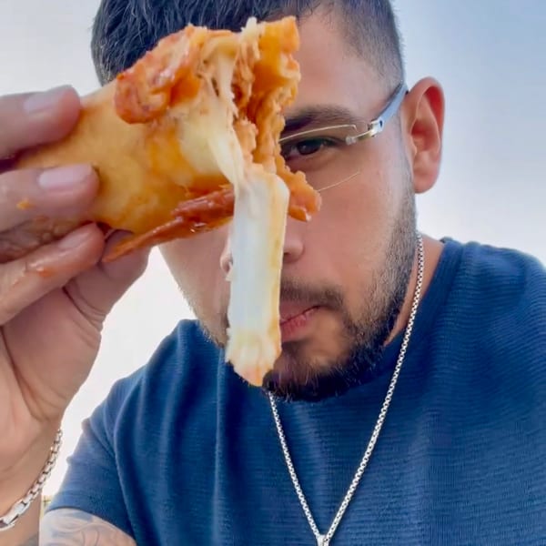 Man in glasses holding a cheesy, saucy empanada with stringy cheese in front of his face.