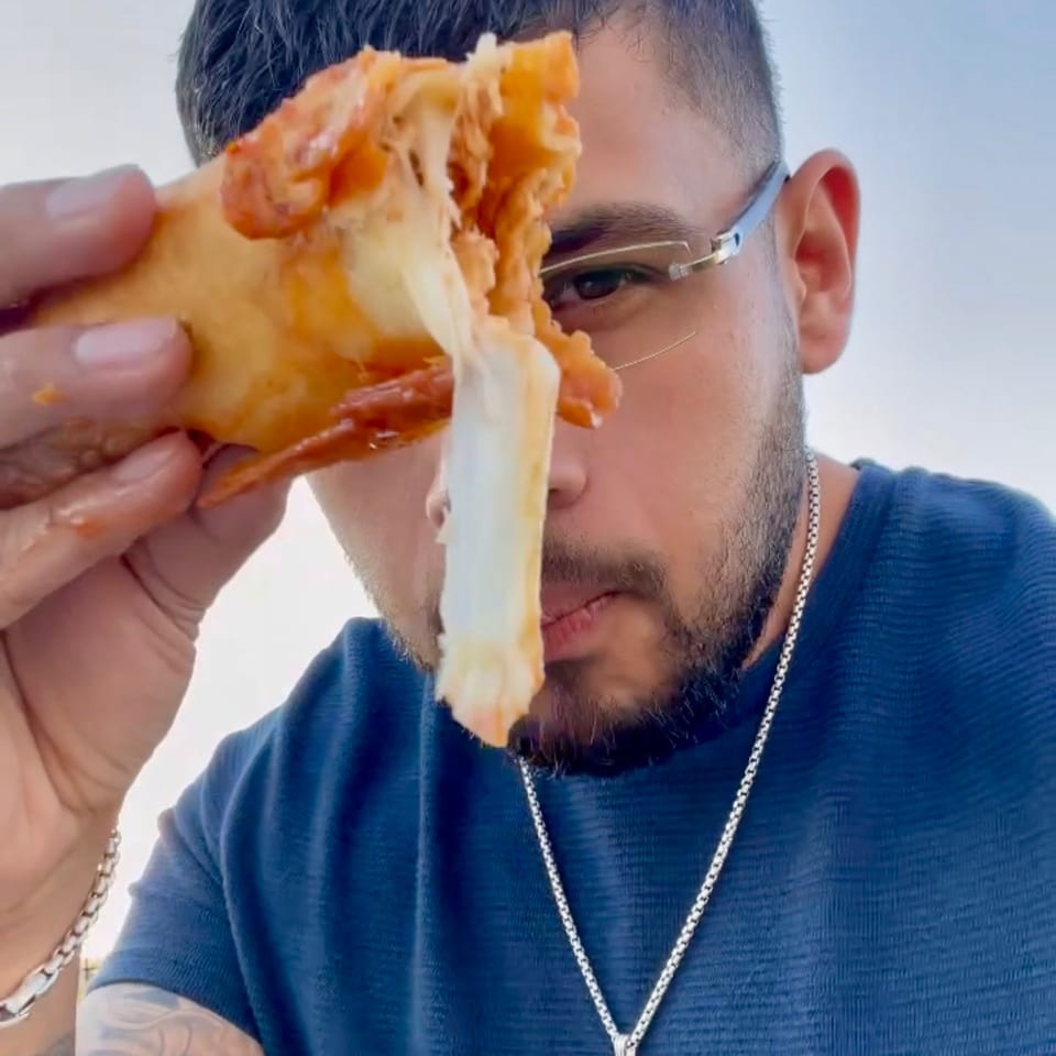 Man in glasses holding a cheesy, saucy empanada with stringy cheese in front of his face.