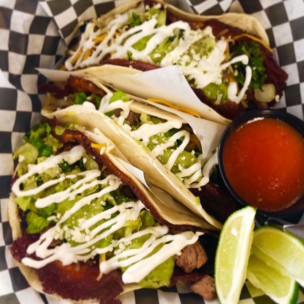 Three El Burro Loco tacos with meat, lettuce, cheese, and crema, with salsa and lime wedges.