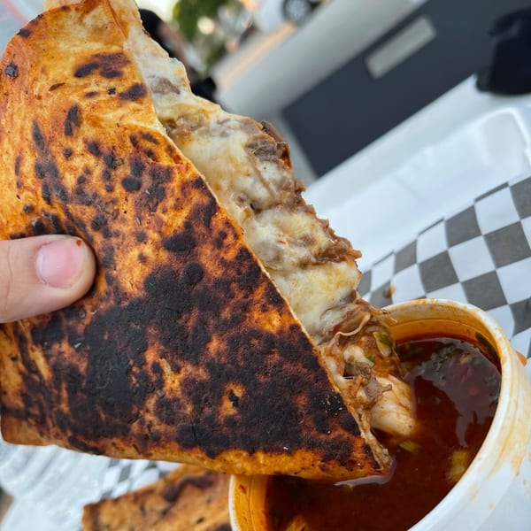 Hand holding a crispy birria quesadilla, dipping it into a cup of rich consommé.