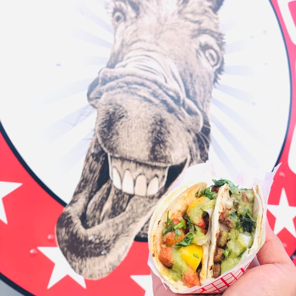 Two tacos with various fillings, held in a hand, against a backdrop of a donkey's head.